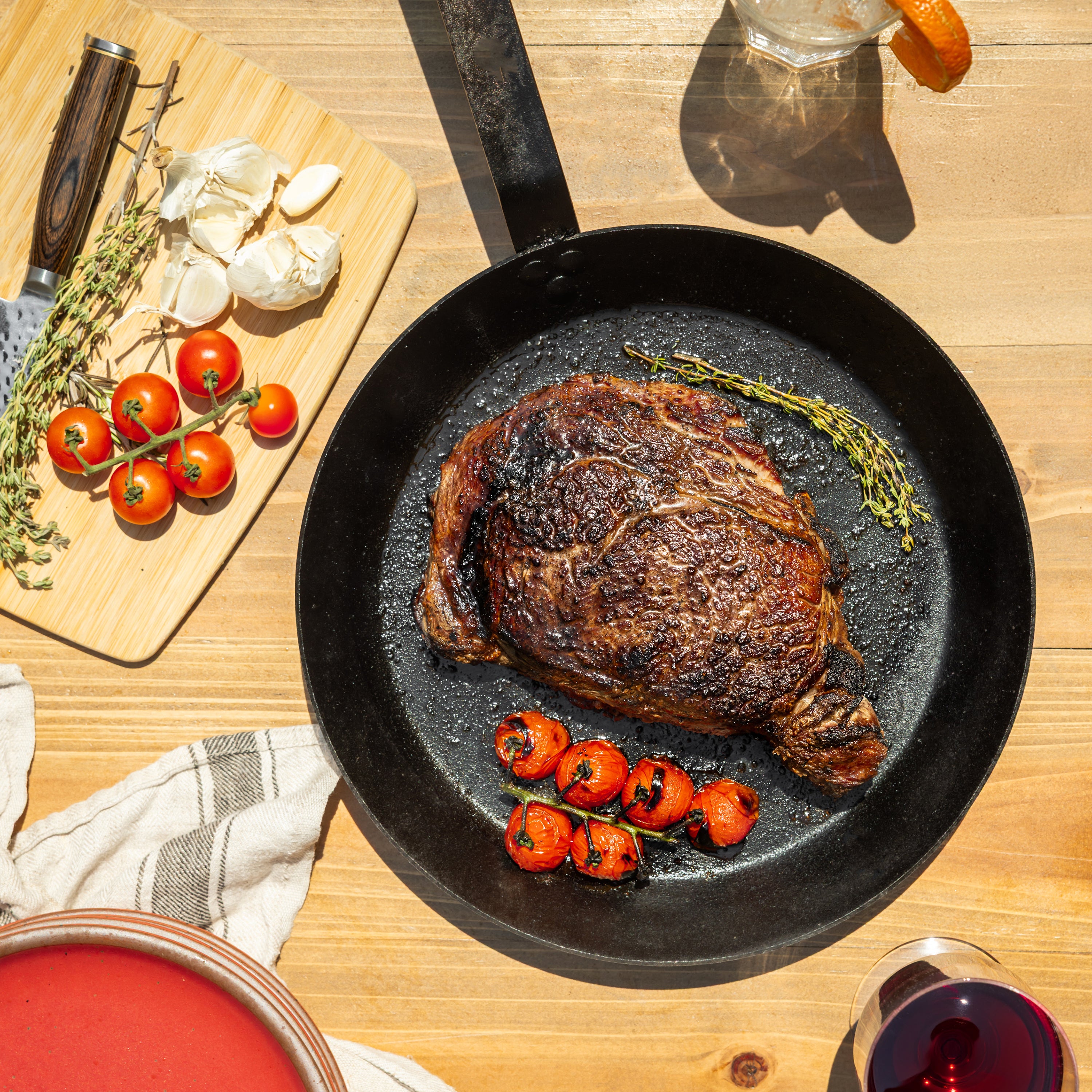 A perfectly seared steak sits in a black carbon steel skillet on a wooden table outside, surrounded by roasted cherry tomatoes, garlic, herbs, a cutting board with tomatoes and garlic, and a glass of red wine.