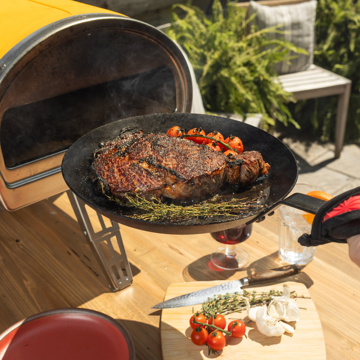 Someone removing skillet from an outdoor pizza oven, revealing a perfectly seared steak.