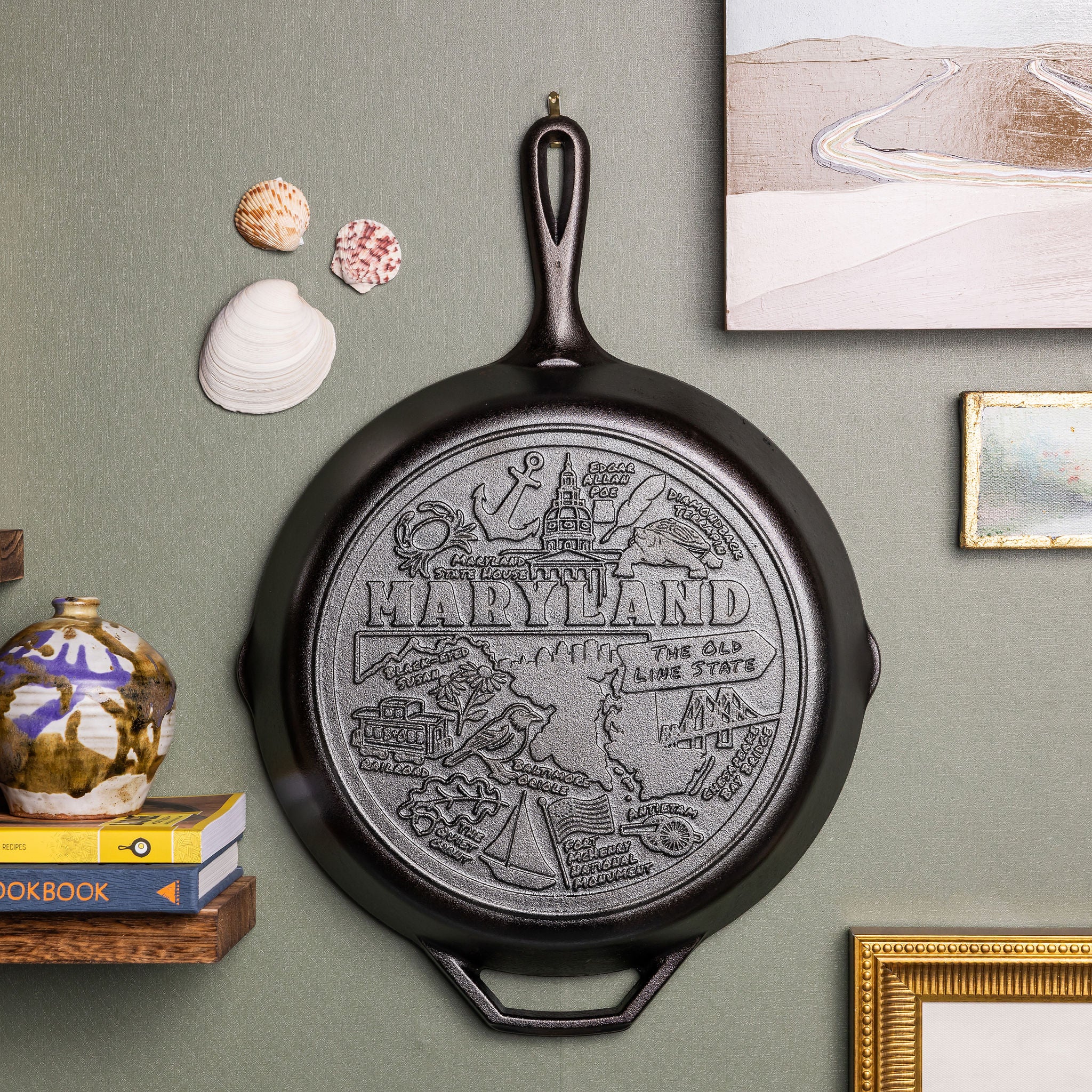 Decorative cast iron skillet with 'Maryland' design on a wall with books and seashells.