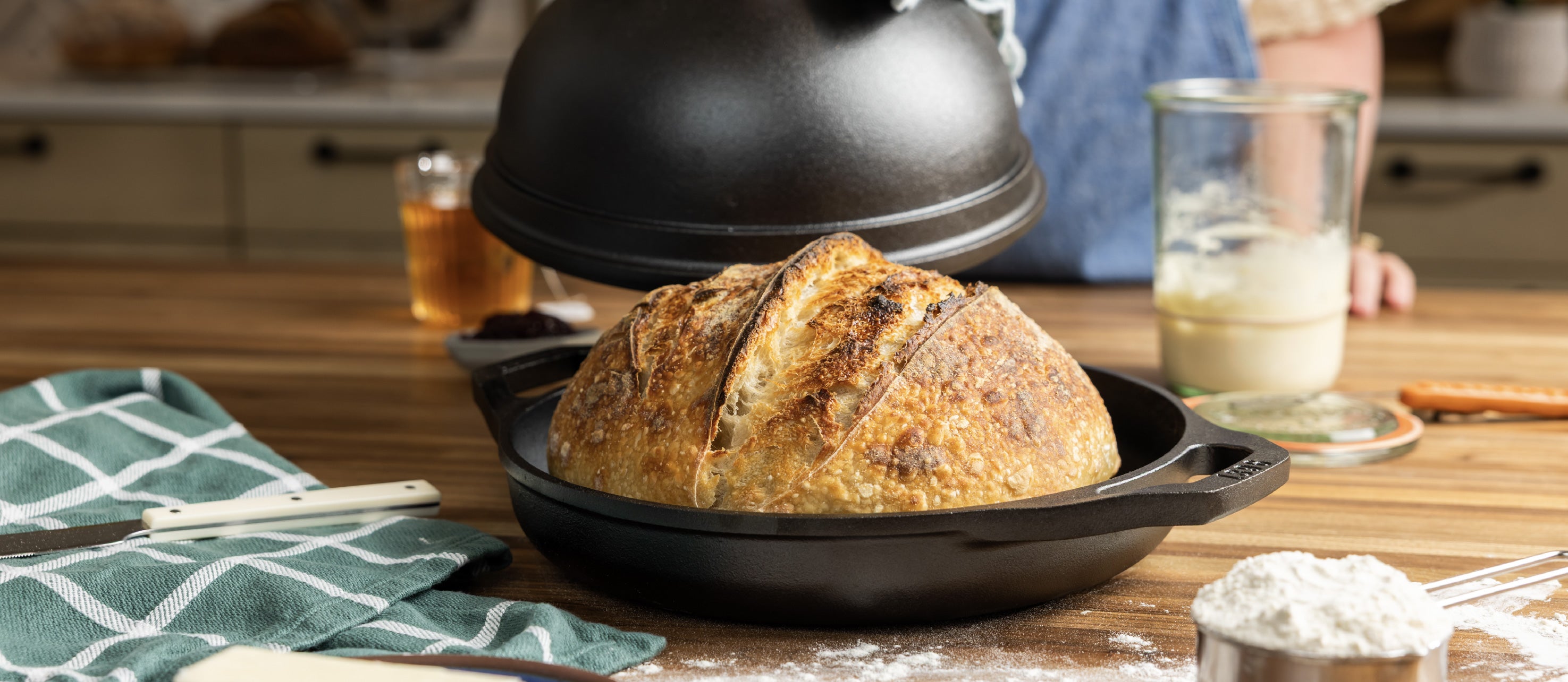 A cast iron bread oven with a lid sits on a wooden kitchen table, with a baked bread loaf inside. A person in a blue shirt is partially visible in the background, along with a glass of milk, a green checkered cloth, and baking ingredients.