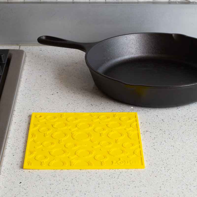 Yellow silicone trivet placed flat on counter next to stove and skillet.