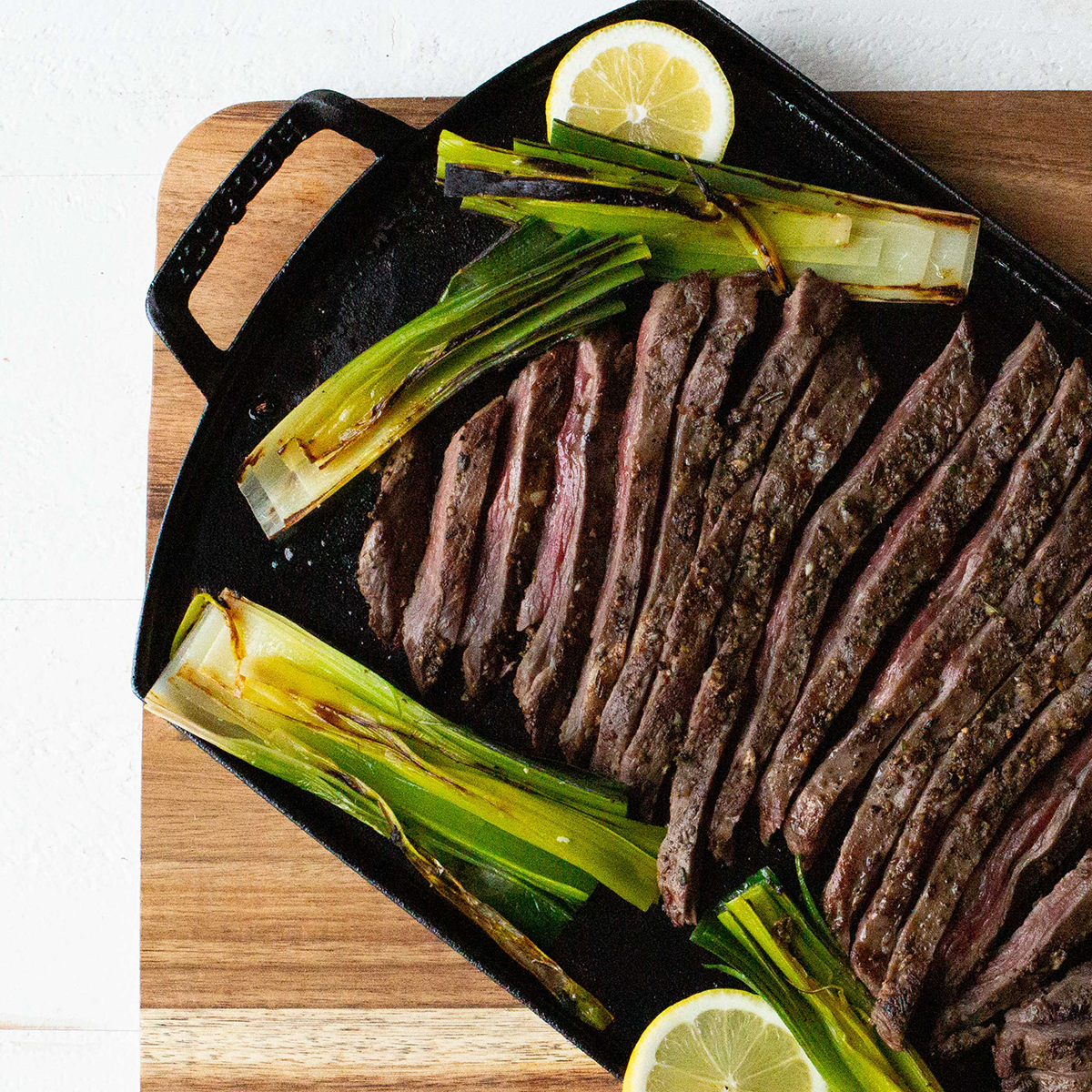 Sliced steak with lemons and green onions on griddle over a wooden board.