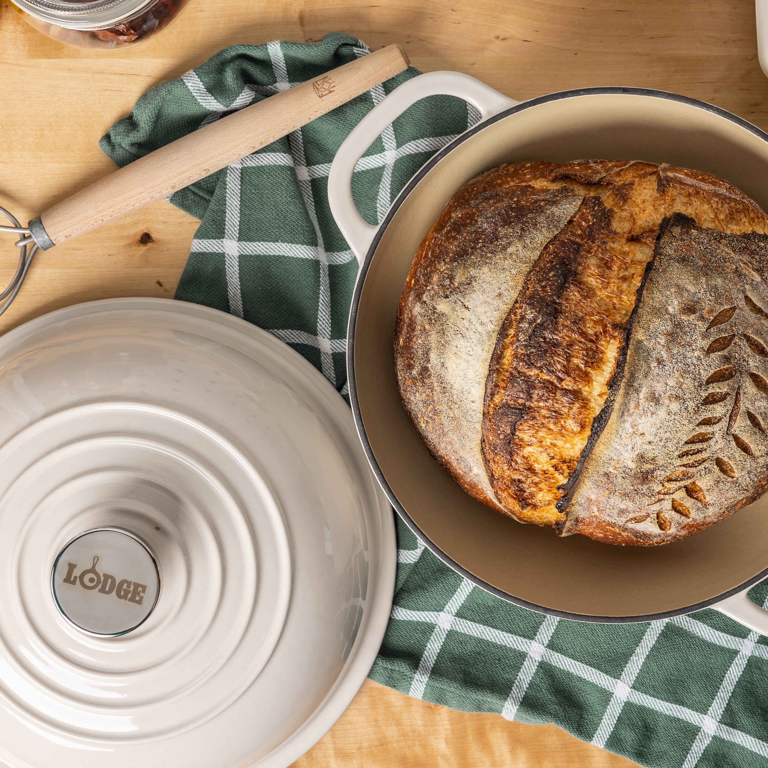 A Lodge enameled cast iron round bread oven with its lid off, showing a rustic artisan sourdough bread inside, styled on a wooden table with a green checkered cloth.