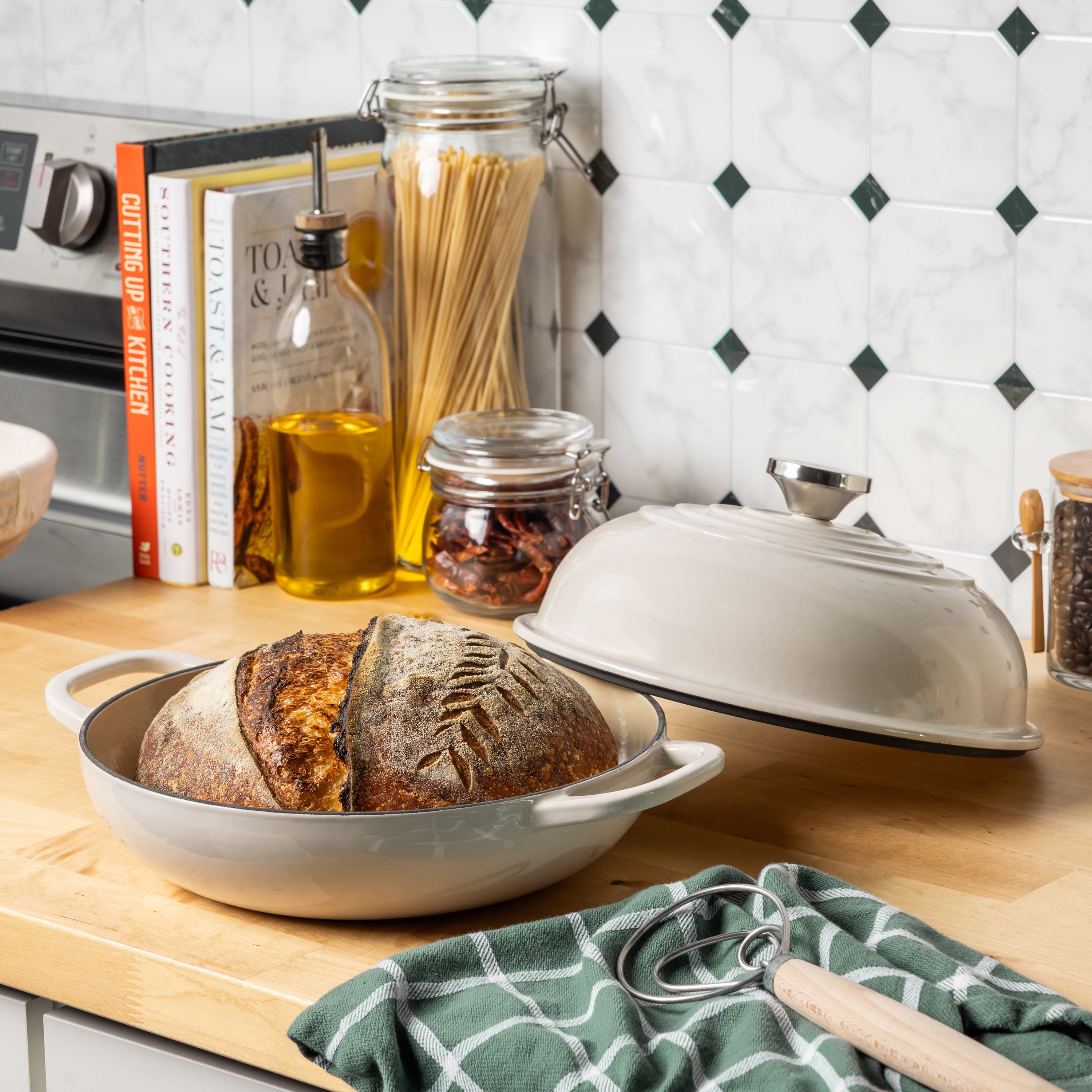An enameled bread baking dish with a rustic crusty loaf of bread inside, shown on a wooden kitchen counter with a matching covered dish, books, jars, and tiled wall in the background.