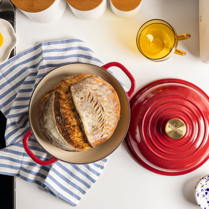 A Lodge enameled cast iron round bread oven with its lid off, showing a rustic artisan sourdough bread inside and a tan interior.