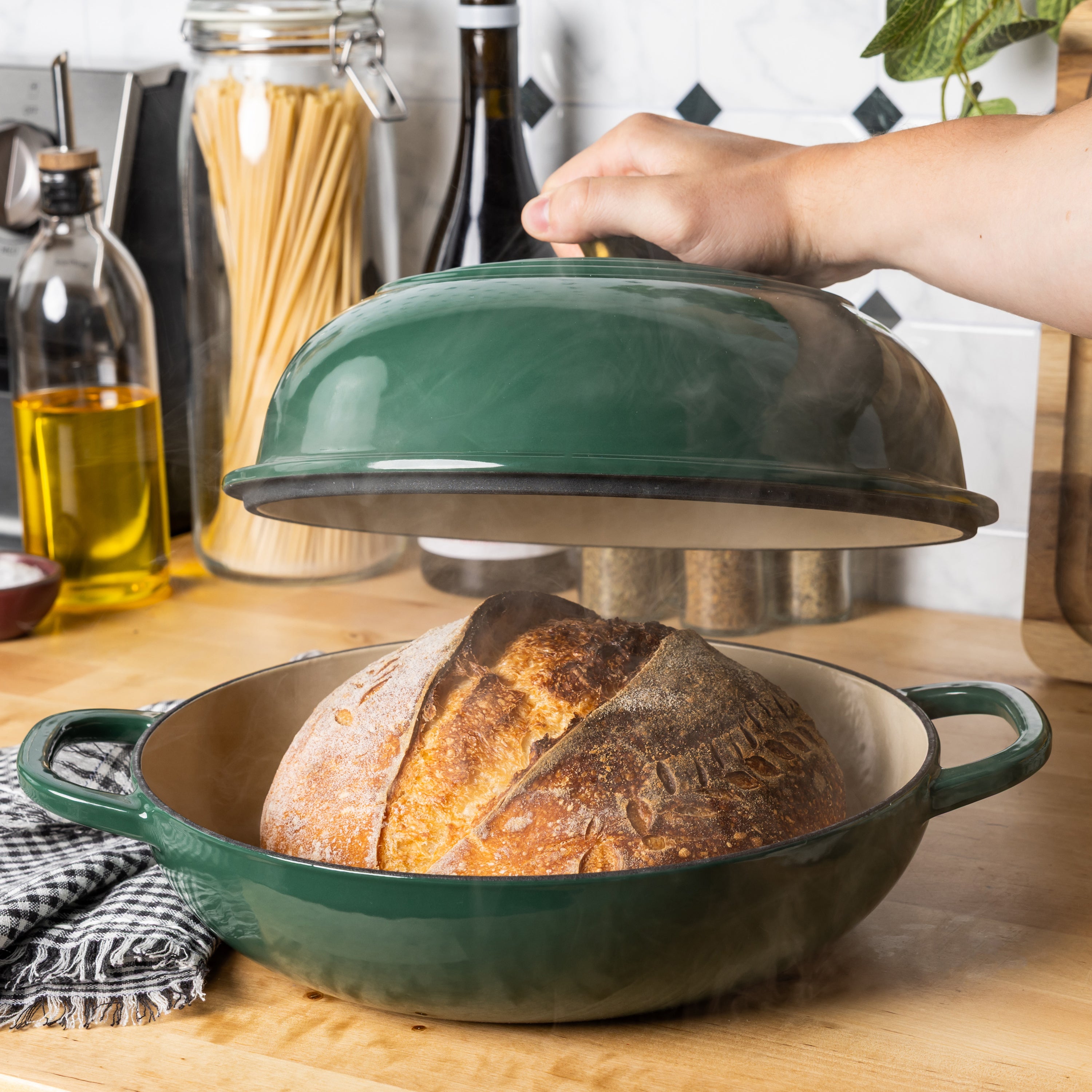 A dark green enameled cast iron bread oven with a matching lid, shown on a wooden countertop with a freshly baked loaf of bread inside. A person's hand is lifting the lid, and kitchen items like oil, pasta, and wine bottles are visible in the background.