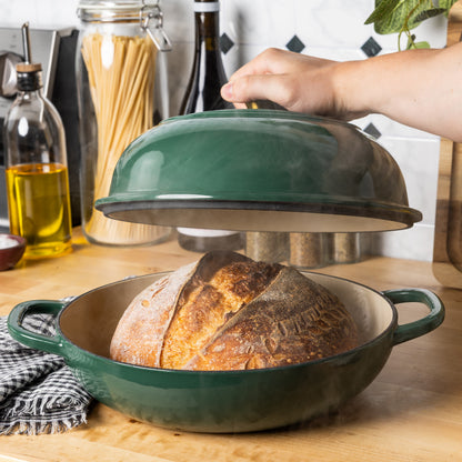 A dark green enameled cast iron bread oven with a matching lid, shown on a wooden countertop with a freshly baked loaf of bread inside. A person's hand is lifting the lid, and kitchen items like oil, pasta, and wine bottles are visible in the background.