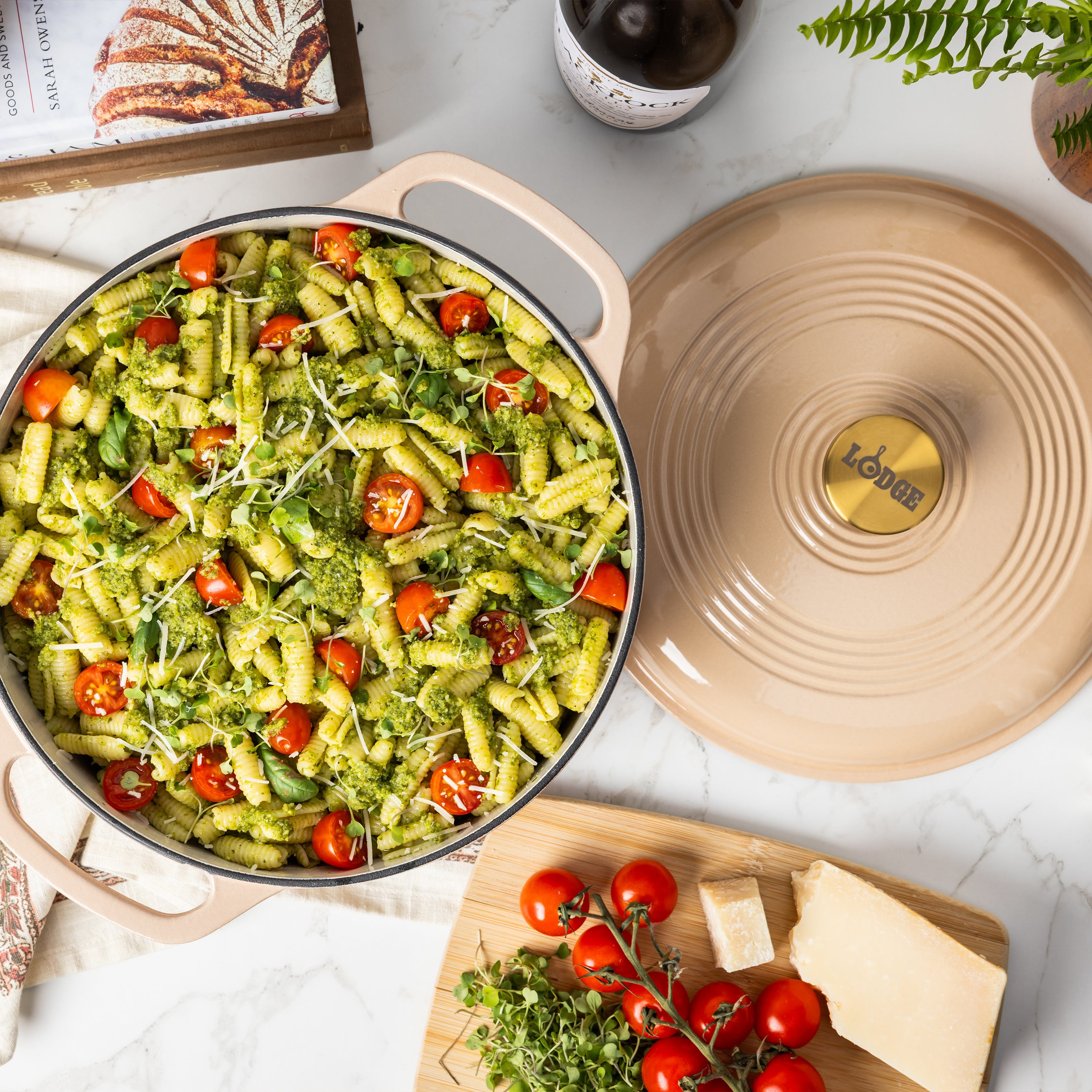 A beige enameled cast iron Dutch oven filled with green pesto pasta, tomatoes, and microgreens, shown on a white marble surface with its matching lid beside it and cooking ingredients nearby.
