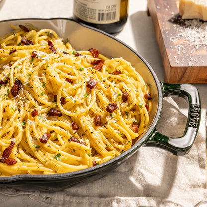 A close-up of creamy carbonara pasta in a dark green cast iron skillet with a black handle, garnished with crispy bacon bits, herbs, and grated cheese. A bottle and a cutting board with cheese are visible in the softly lit background.