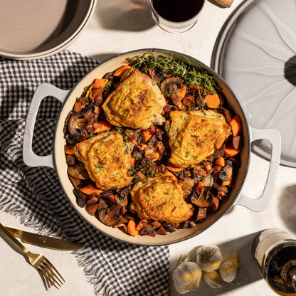 A top-down view of a white enameled cast iron Dutch oven filled with roasted chicken thighs, carrots, mushrooms, and herbs, styled on a white surface with a black-and-white checkered cloth, gold-toned cutlery, and a glass of red wine.