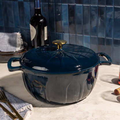 A dark blue enameled cast iron Dutch oven with a lid and gold-tone knob, set on a light stone countertop with a blue tiled backsplash, a wine bottle, and dining props in the background.