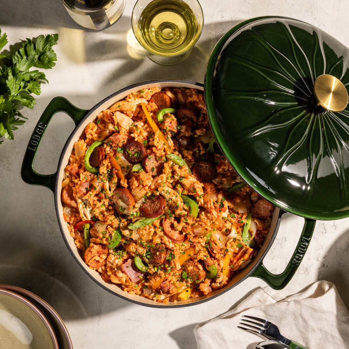A top-down view of a green enameled cast iron Dutch oven filled with a sausage and rice dish, with its matching lid resting beside it on a light stone surface. The brand name 'LODGE' is visible on the side of the pot.