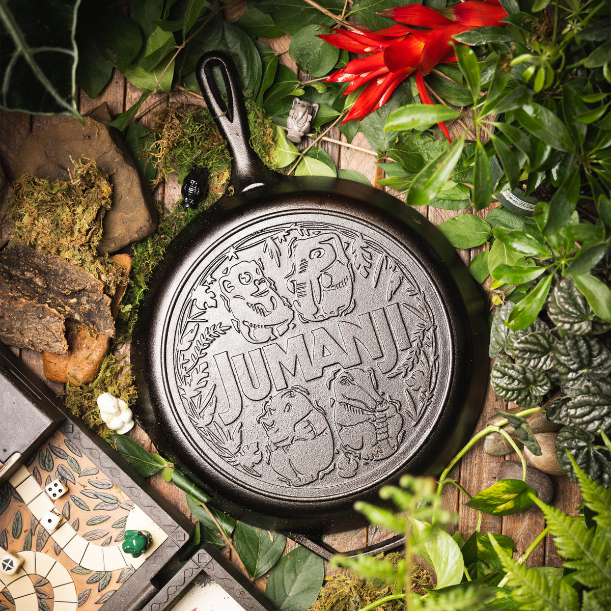 A cast iron skillet with an embossed 'JUMANA' design, surrounded by lush green foliage, moss, rocks, and a small game board on a rustic wooden surface.