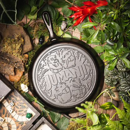 A cast iron skillet with an embossed 'JUMANA' design, surrounded by lush green foliage, moss, rocks, and a small game board on a rustic wooden surface.