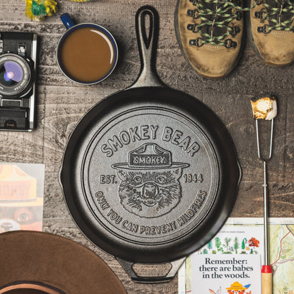 Cast iron skillet with Smokey Bear logo on a rustic wooden surface with coffee, boots, and a camera.