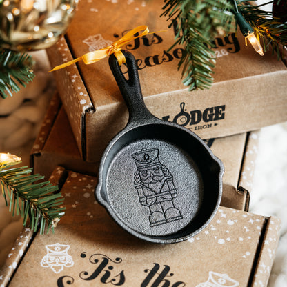 A small black cast-iron nutcracker ornament shaped like a miniature skillet, displayed on top of brown gift boxes with holiday greenery and warm string lights around it.