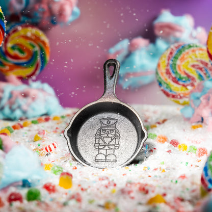 A small skillet with a nutcracker engraved in the center, placed on white candy-coated sugar with colorful sprinkles. The background is a soft-focus pink and purple with blurred lollipops and pastel candy pieces.
