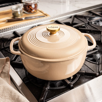 A Lodge enameled cast iron Dutch oven with lid in a warm beige color, sitting on a black gas stovetop in a kitchen setting.