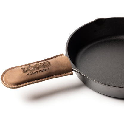 A close-up studio shot of a cast iron skillet with a tan leather handle cover embossed with the brand name 'LODGE CAST IRON' on a white background.