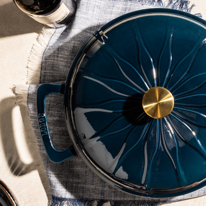 A dark teal blue enameled cast iron Dutch oven with a matching lid featuring a starburst pattern and a gold knob, styled on a blue fringed placemat with a bottle partially visible.