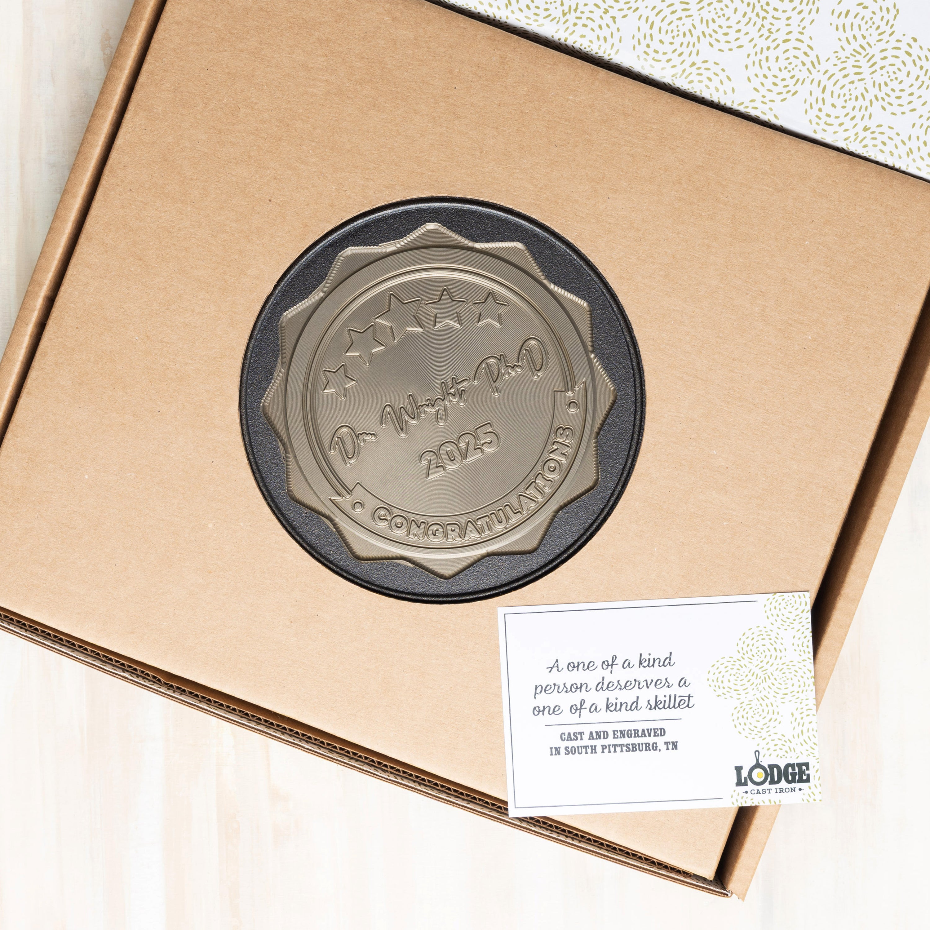 Bronze engraved skillet in a cardboard box with a card attached, on a white background.