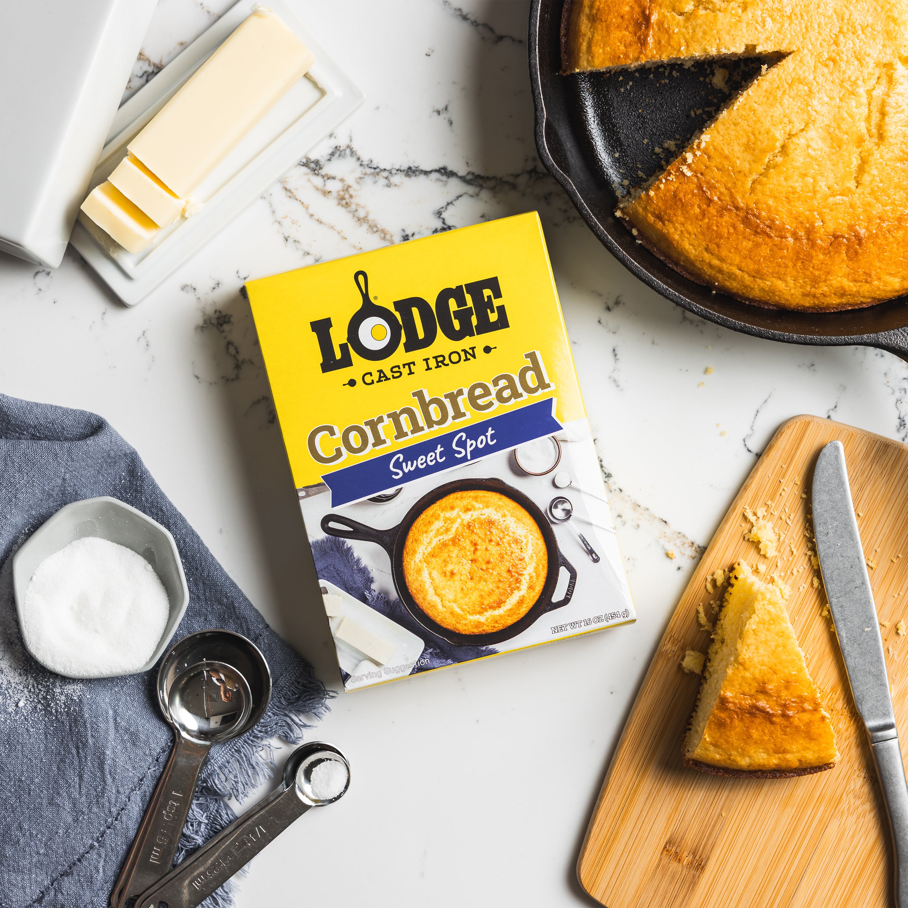 Lodge cornbread mix box with cornbread on a cutting board, knife, and measuring cups on a marble surface.