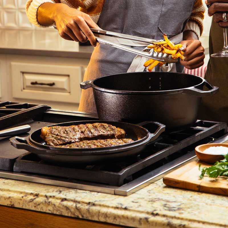 Someone frying French Fries in dutch oven on stove while steak is cooking in lid portion on a different burner.