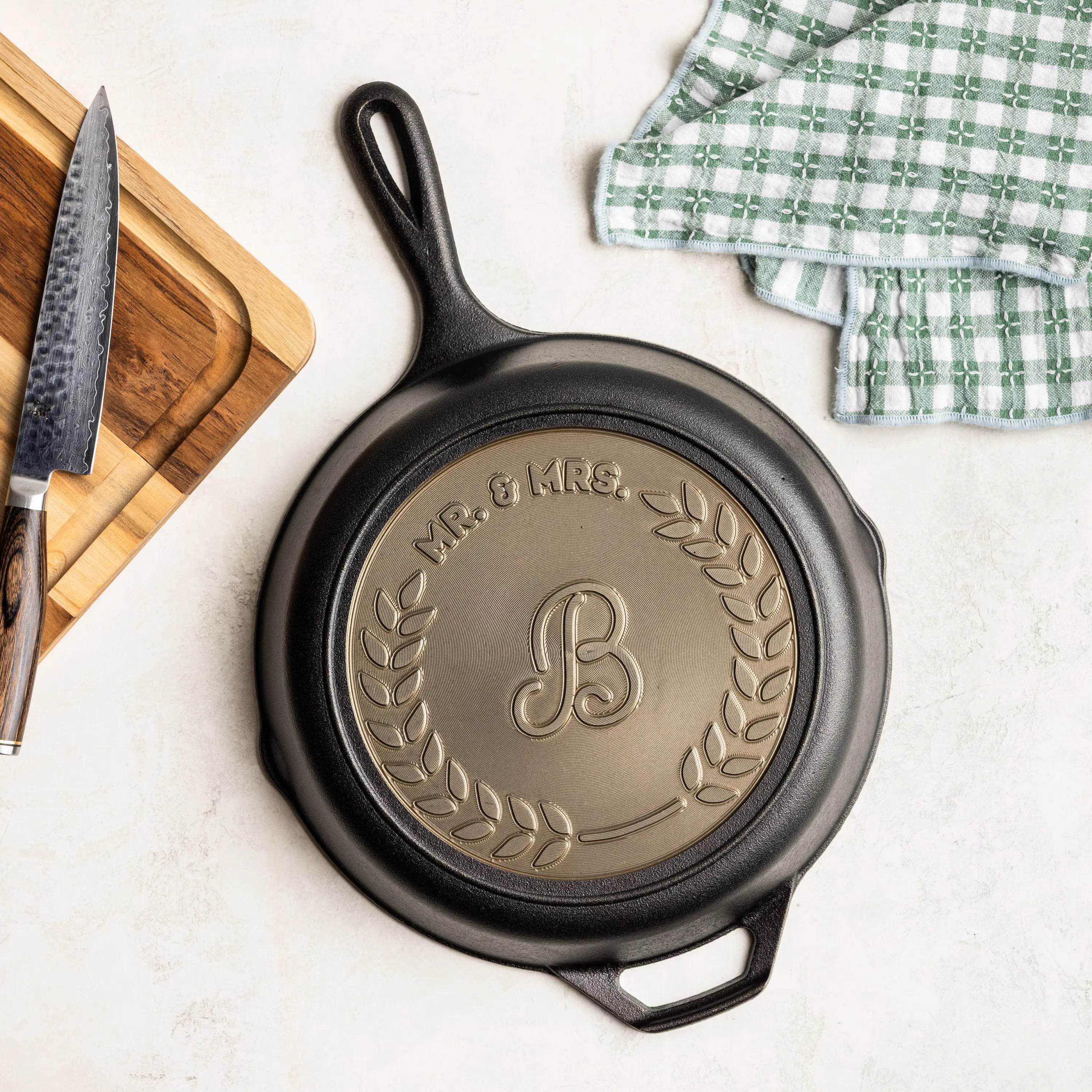 Black cast iron skillet with 'Mr. & Mrs.' and a 'B' emblem on a white surface with a knife and towel.
