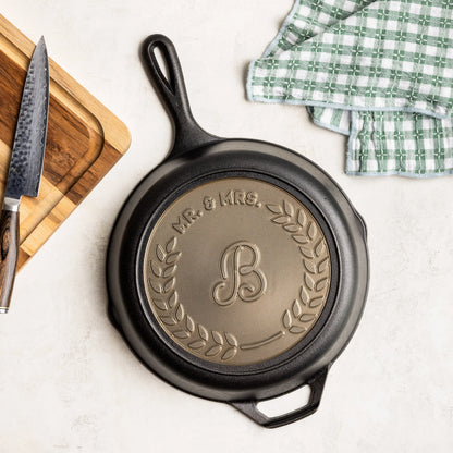 Black cast iron skillet with 'Mr. & Mrs.' and a 'B' emblem on a white surface with a knife and towel.