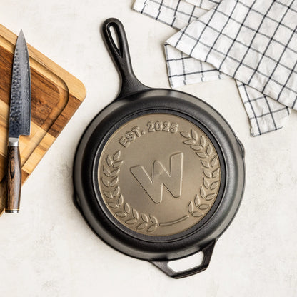 Black cast iron skillet with engraved design on a white surface with a knife and cutting board.