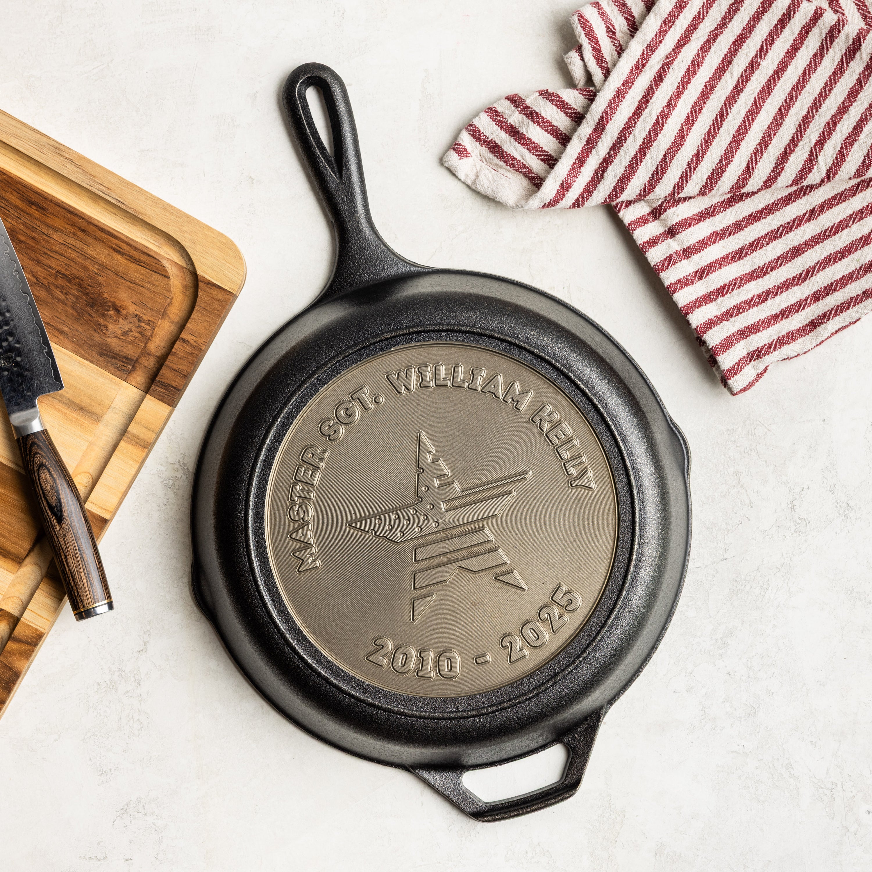 Black cast iron skillet with engraved design on a white surface with a wooden cutting board and red and white striped towel.