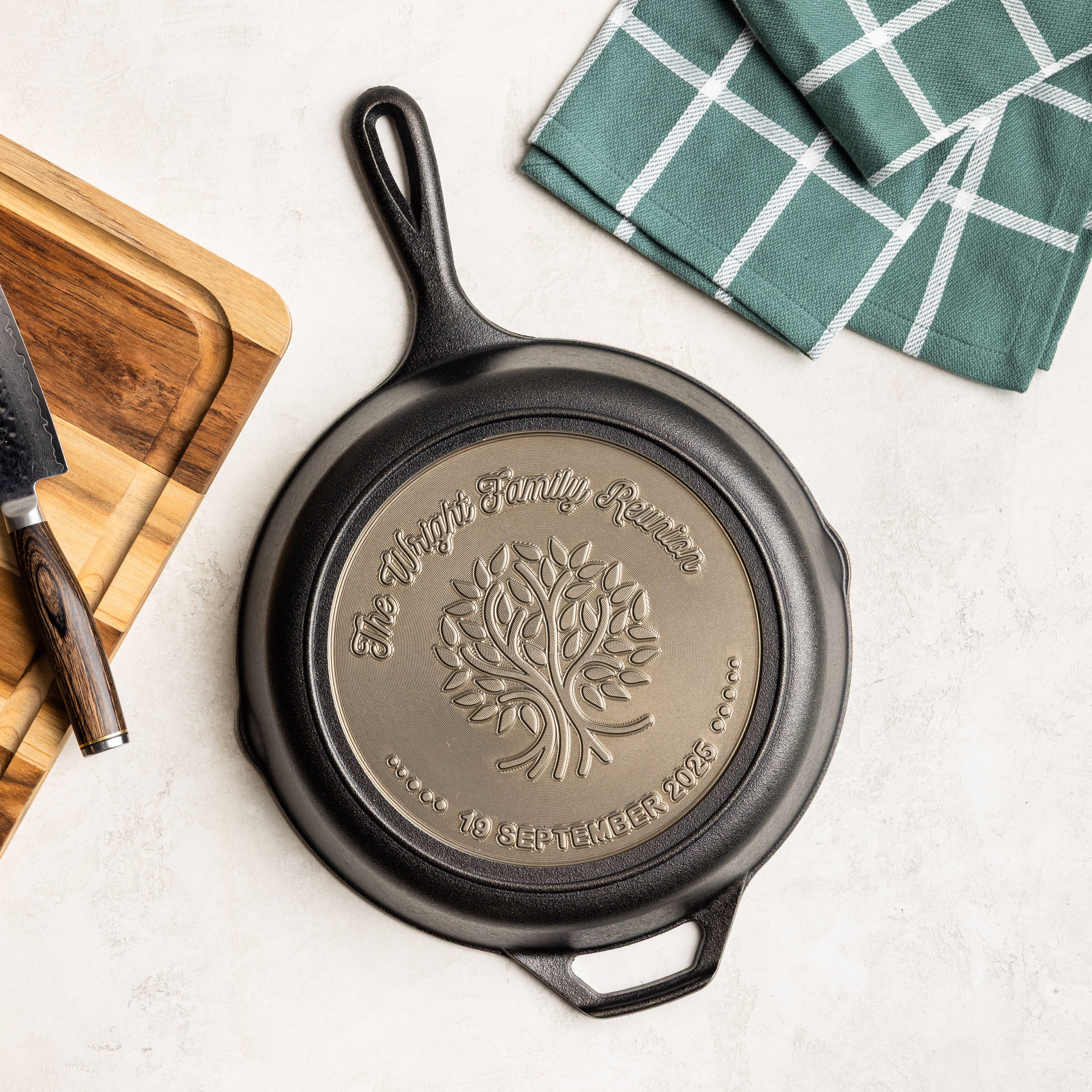 Cast iron skillet with engraved design on a white surface with a wooden cutting board and green checkered towel.