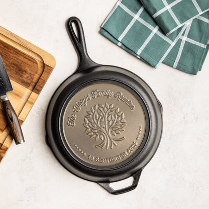 Cast iron skillet with engraved design on a white surface with a wooden cutting board and green checkered towel.