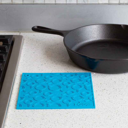 Turquoise silicone trivet placed flat on counter next to stove and skillet.
