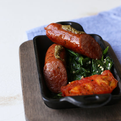 Two sausages with pesto, spinach, and cherry tomatoes in a serve dish on a wooden underliner.