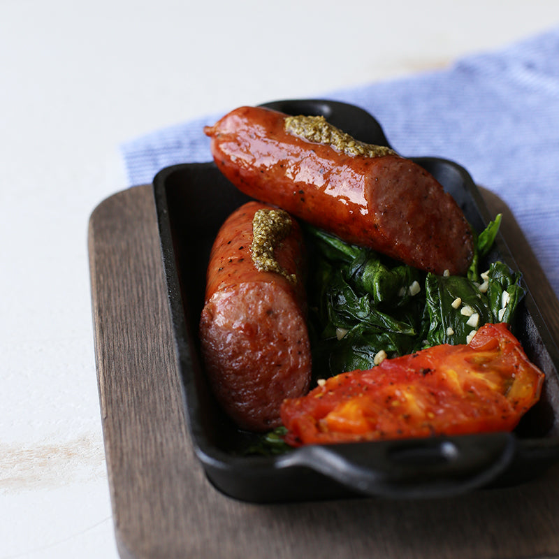Two sausages with pesto, spinach, and cherry tomatoes in a serve dish on a wooden underliner.