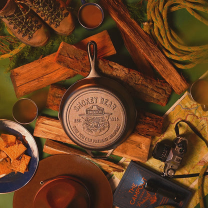 Smokey bear skillet on fire wood surrounded by camping gear.