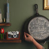 Close up view of the engraved design on bottom of skillet, while skillet is displayed on wall.