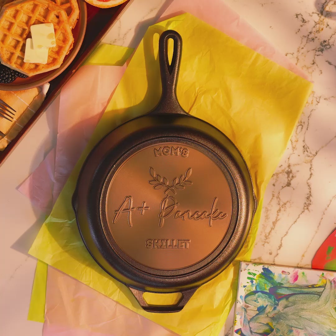 Close-up view of a custom skillet placed near tissue paper and flowers including text "Mom's A+ Pancake Skillet" in editable fields.